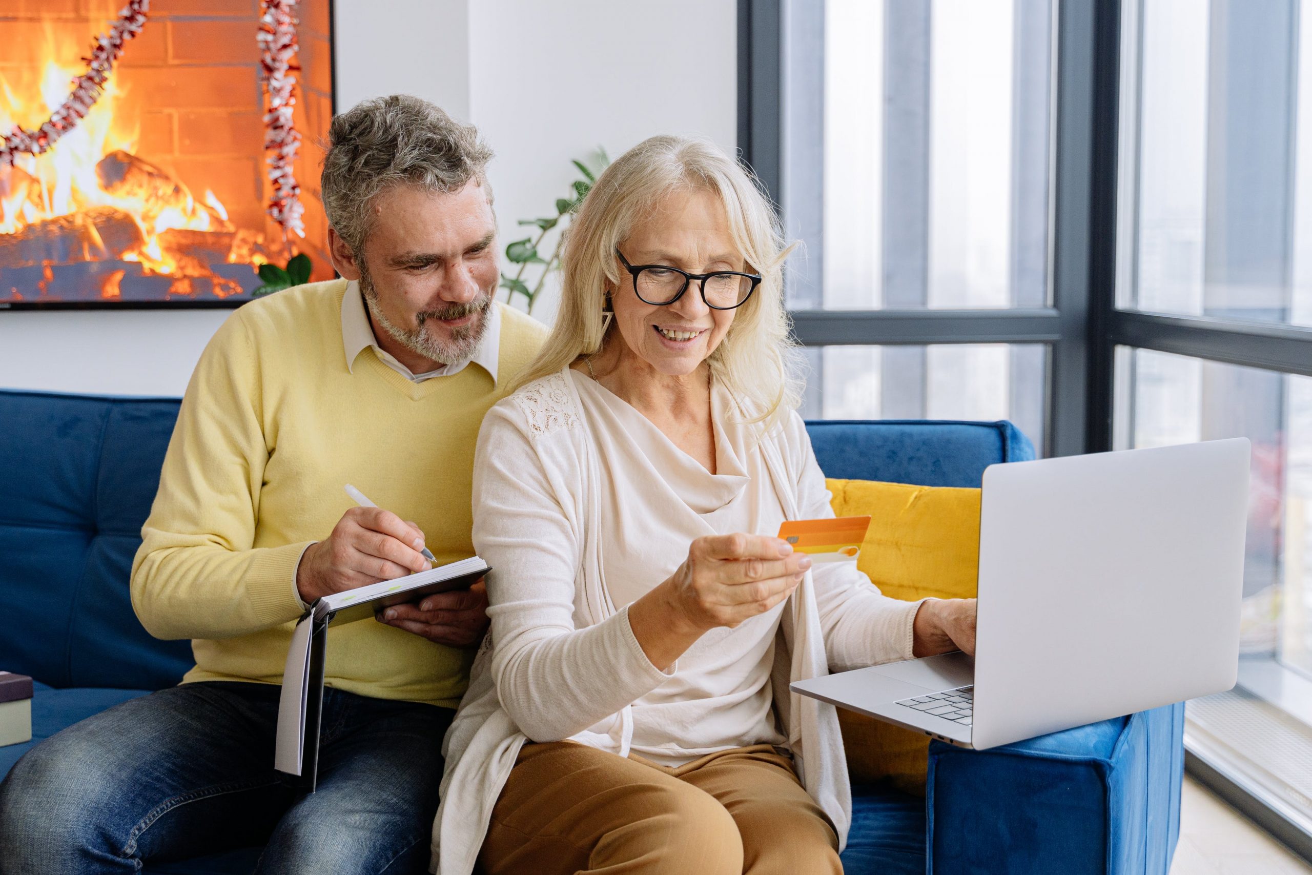 An older couple paying on a laptop using their credit card and writing down the payment in a notebook.