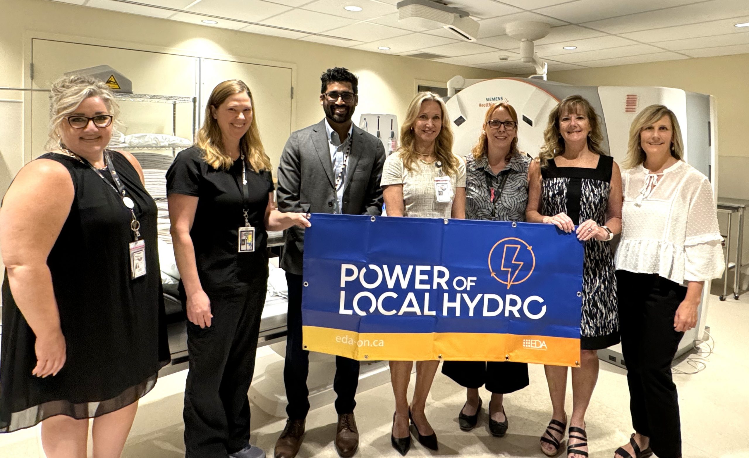 The GrandBridge Energy team standing in front of the new CT Scanner they contributed to the Community while holding a sign that says Power of Local Hydro.